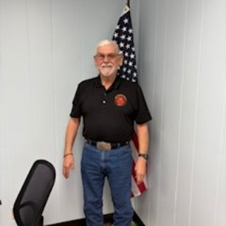 Man standing indoors beside an American flag, wearing a black polo shirt and jeans.
