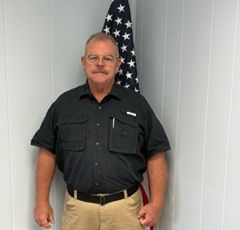 Man standing in front of a U.S. flag, wearing a black shirt and tan pants.