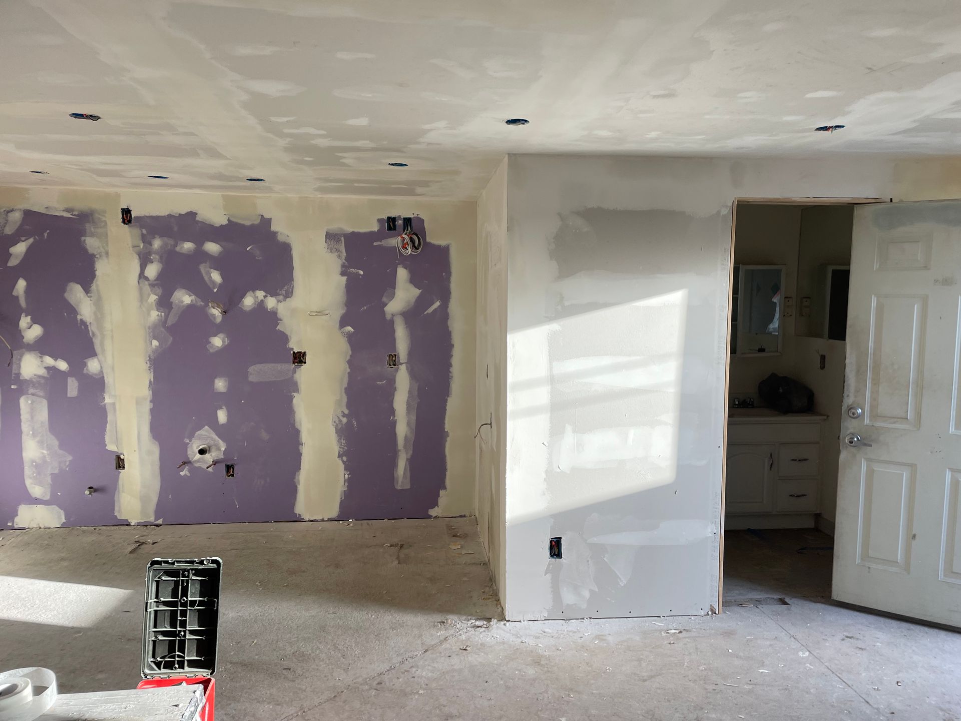 Room under renovation with purple drywall patches, white walls, and an open door to a bathroom.