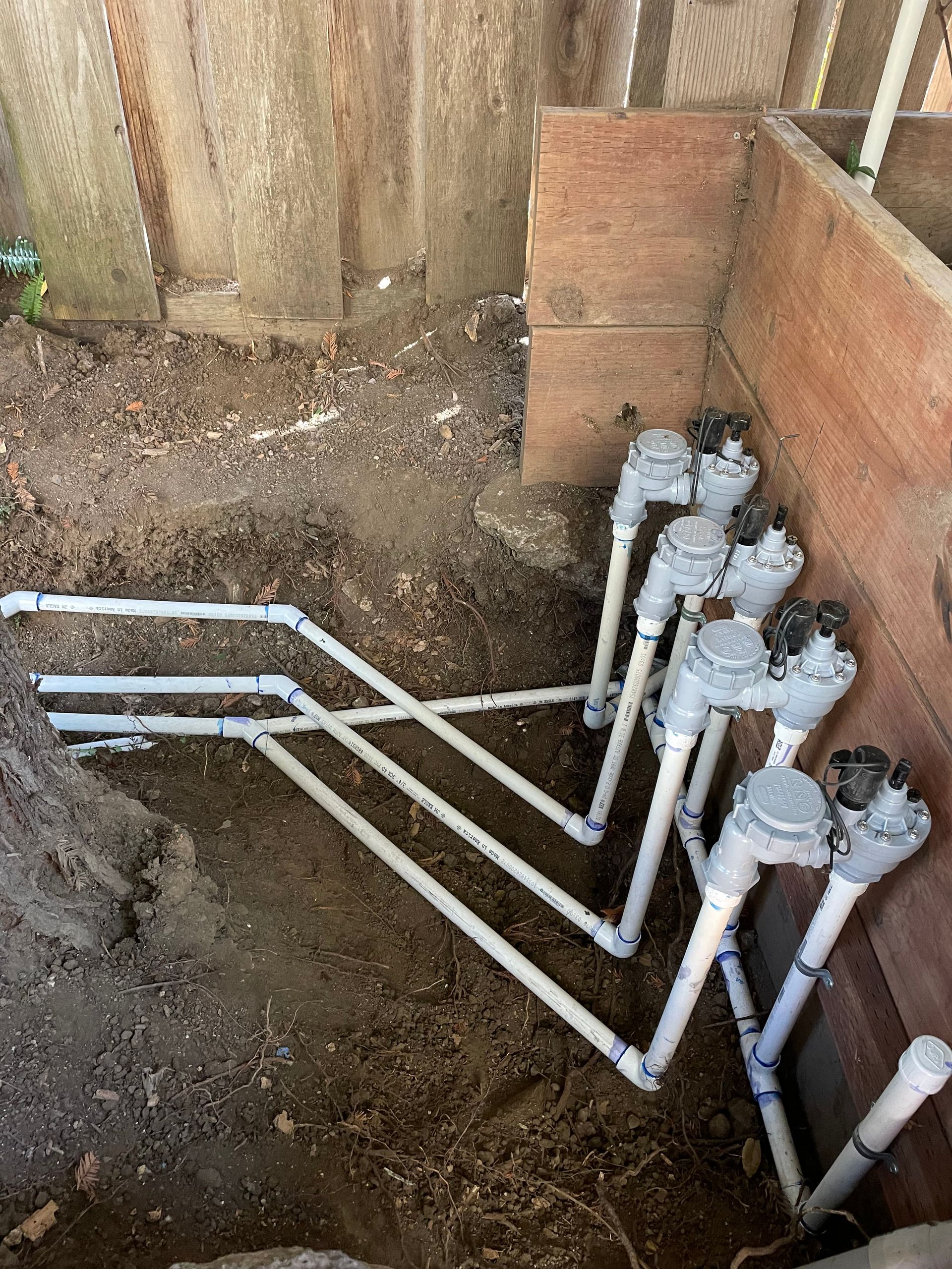 Exposed white PVC pipes in a dirt trench beside a wooden fence, with several capped vertical pipes.