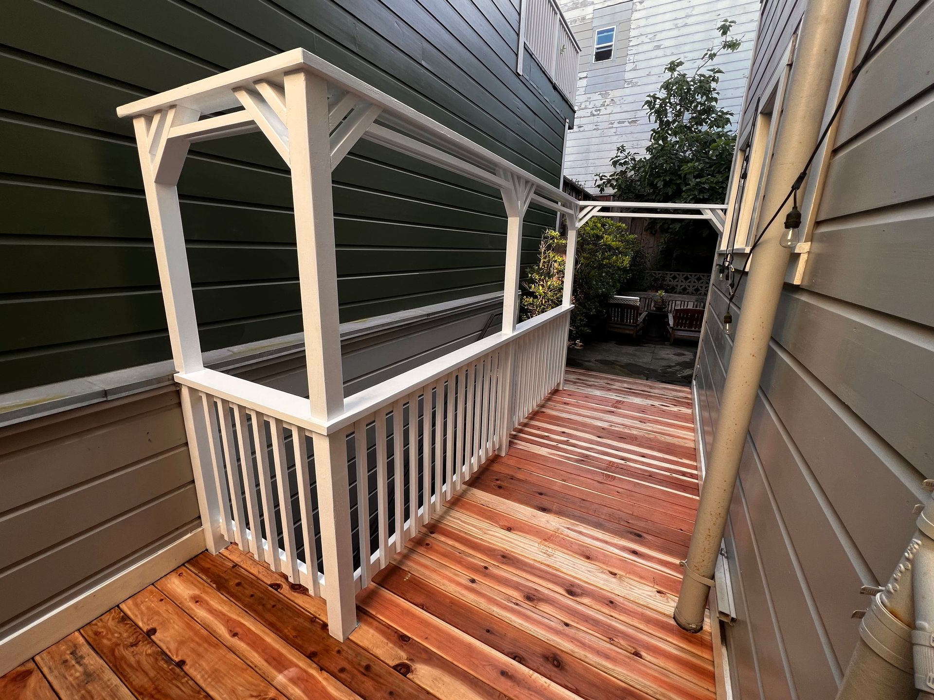 Narrow wooden deck walkway between house walls, with white railing and overhead pergola frame.