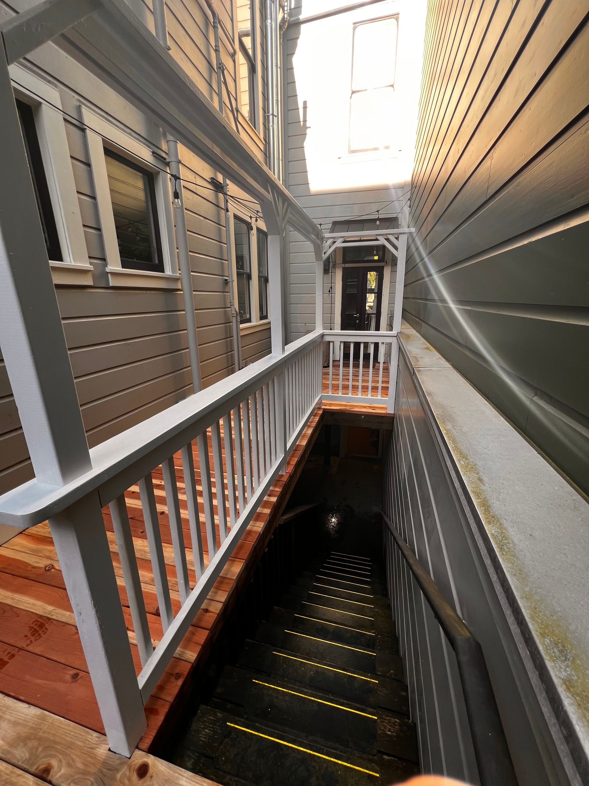 Narrow outdoor stairwell between buildings with white railings and gray siding, lit by sunlight at the top.