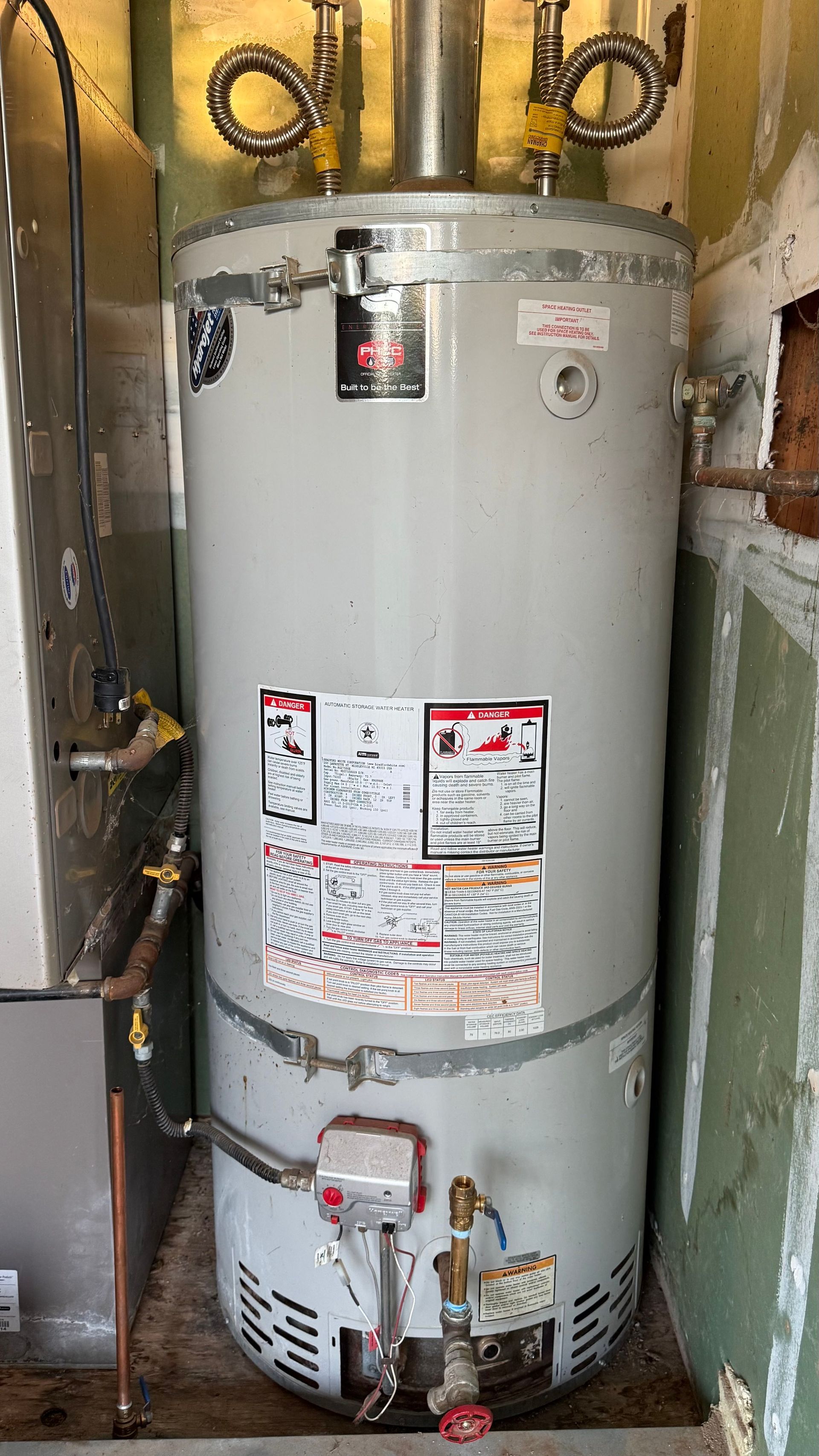 Gray water heater in a utility closet with pipes, valves, and warning labels.