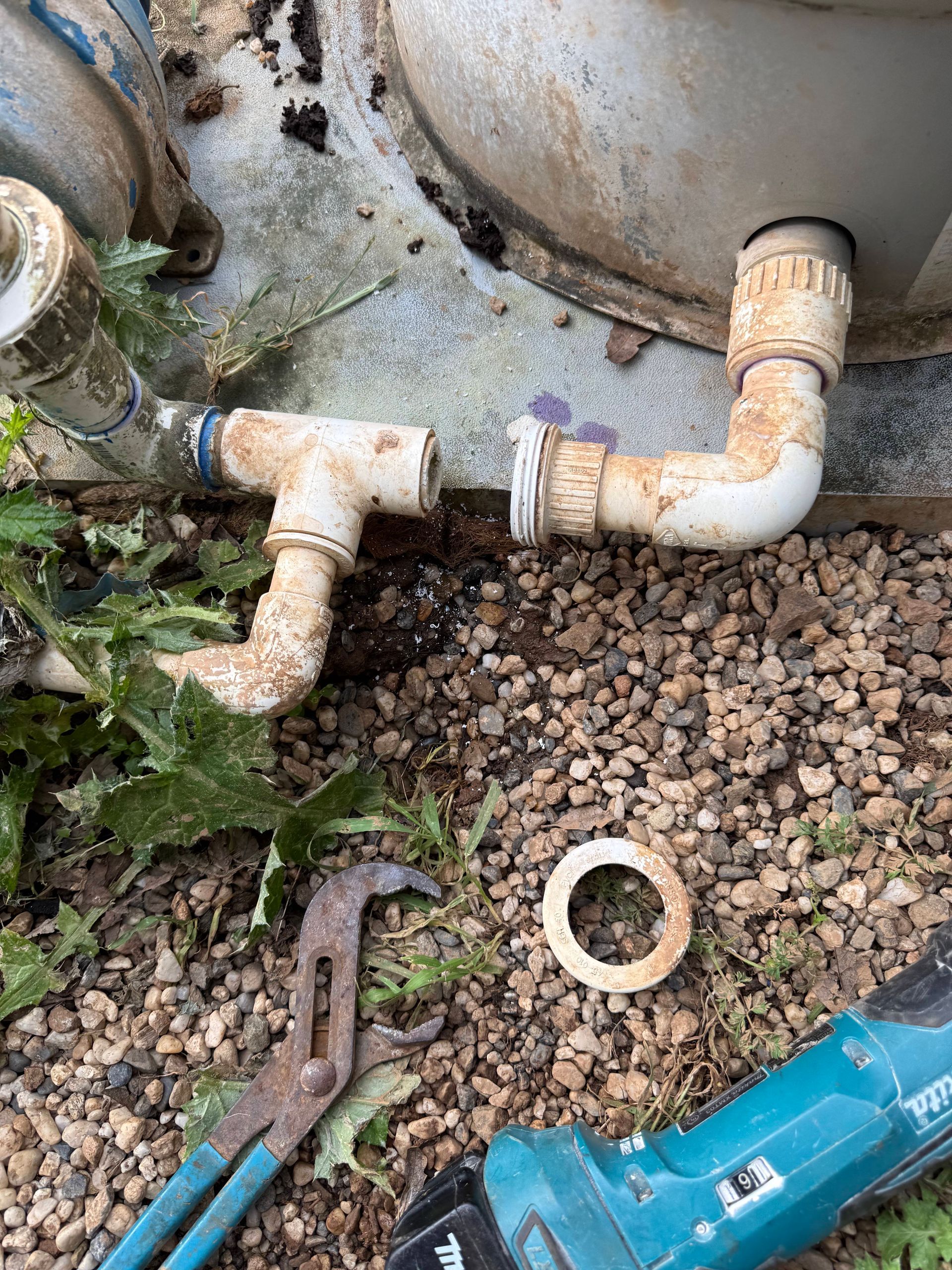 Old outdoor plumbing pipes beside a gravel bed, with a wrench and blue power tool on the ground.