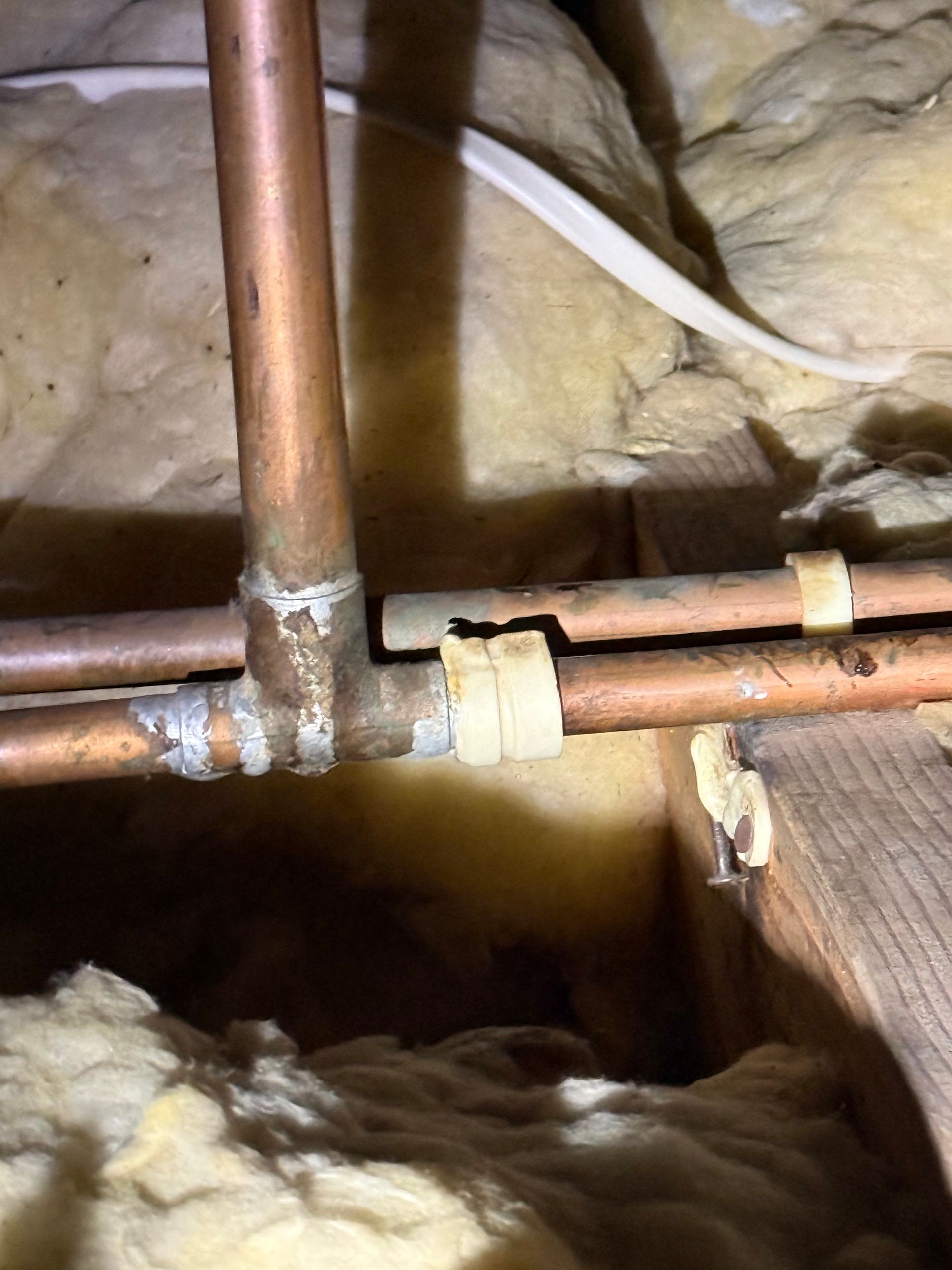 Copper pipes and insulation inside an attic, with a small white clamp on the horizontal pipe.