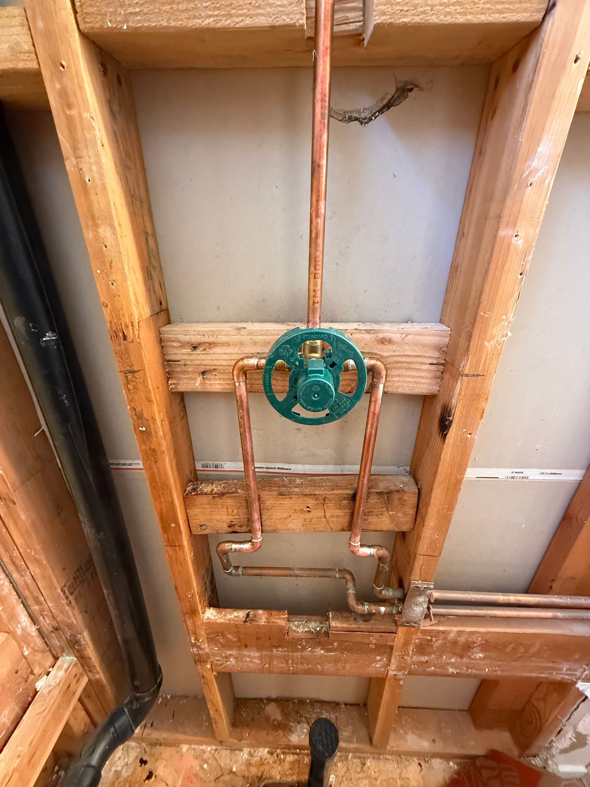 Plumbing pipes and a green valve mounted between exposed wooden wall studs in a utility area