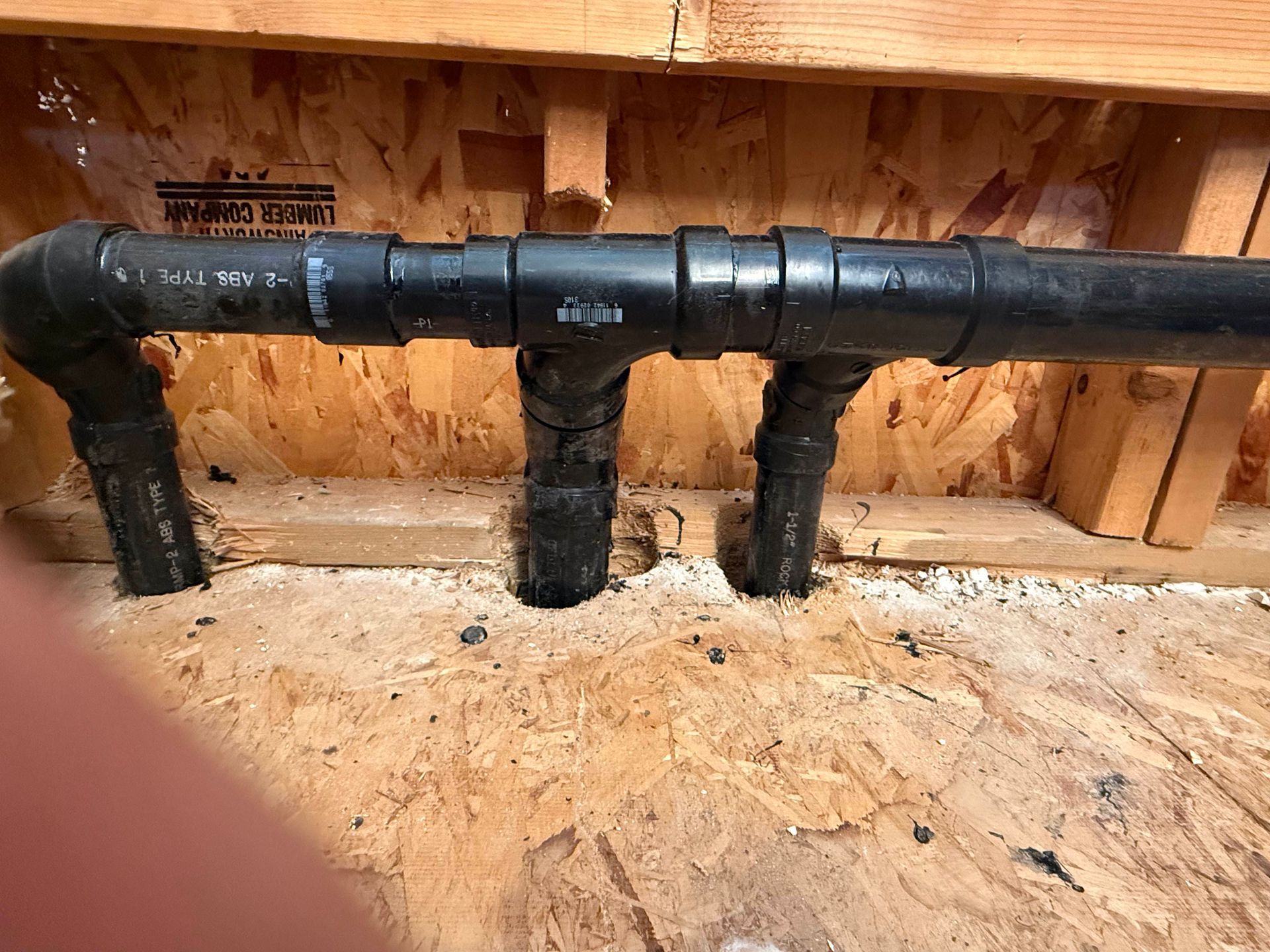 Black PVC plumbing pipes with three downward branches inside a wooden-framed wall cavity.