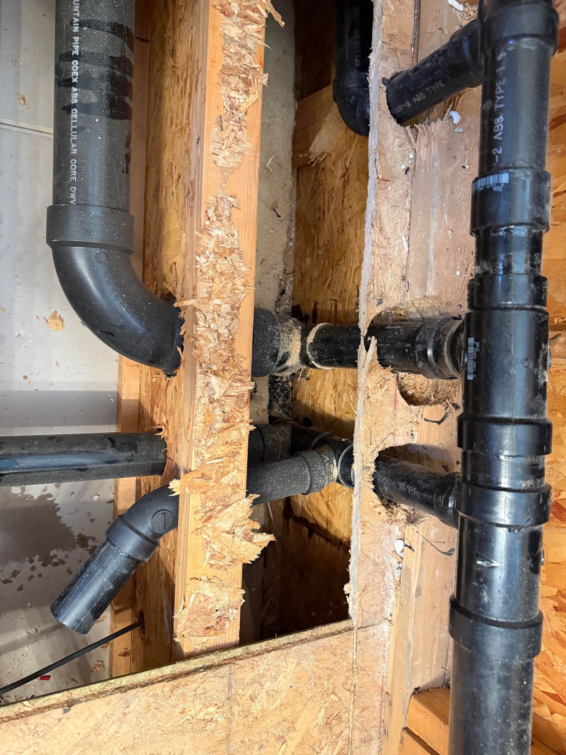 Open wall showing black plumbing pipes, a vertical drain stack, and exposed wooden studs with water damage.