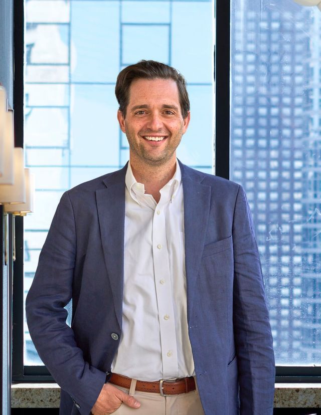 Man in blue blazer and tan pants smiles, standing near a window with city views.