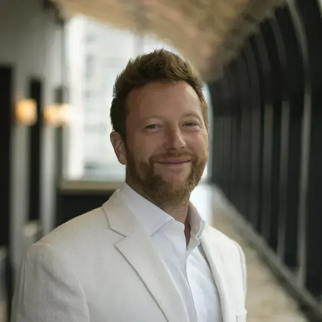 Man in a white blazer and shirt smiling, in a modern hallway with natural light.