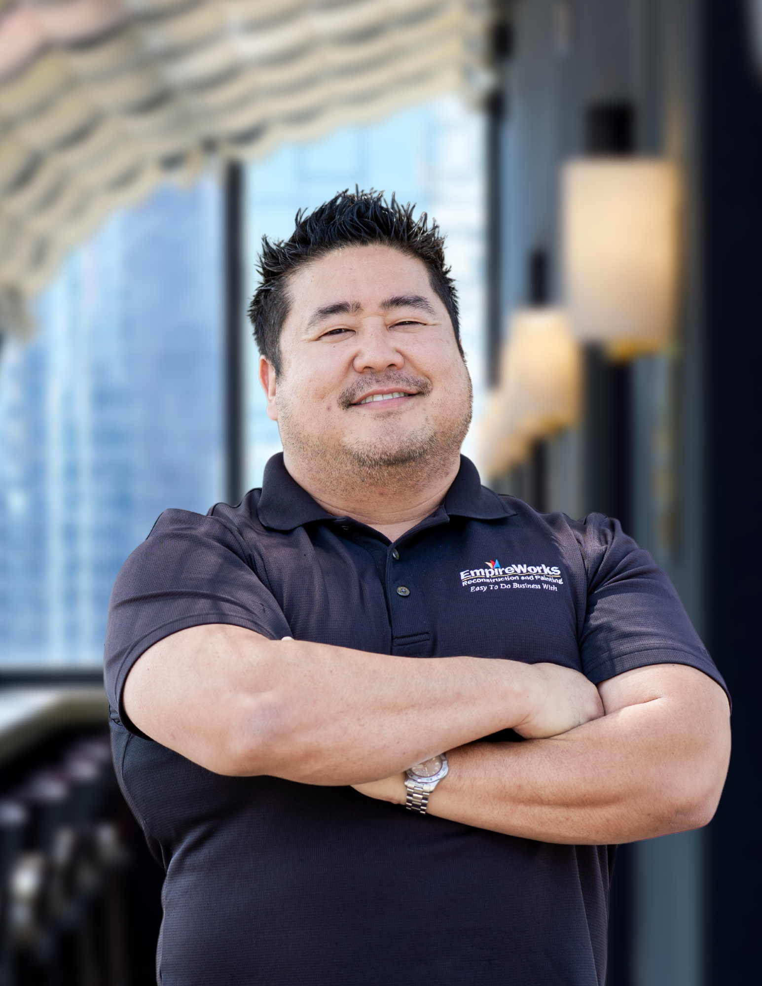 Man with arms crossed, smiling, wearing a black shirt with a logo, in an indoor setting.