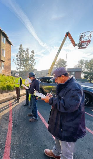 Three people in parking lot, inspecting building; one in lift, one wearing a high-visibility vest. Blue sky.
