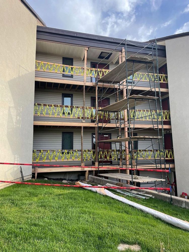 Scaffolding next to a multi-story building with wooden balconies and green railings, grassy area in foreground.