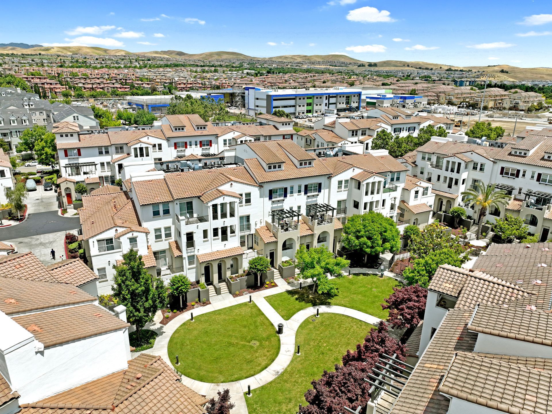 Aerial view of white townhouses with red-tiled roofs, a central green lawn, and a distant cityscape under a blue sky.