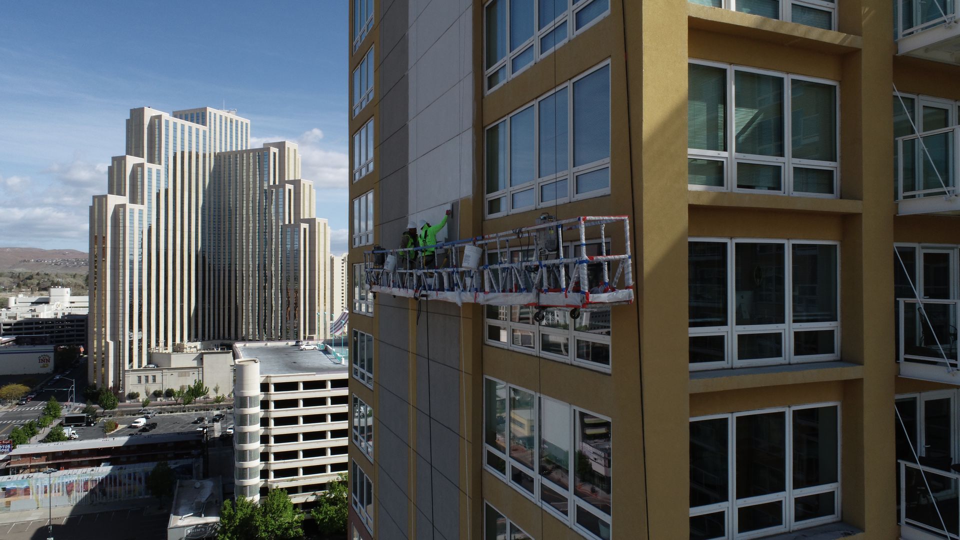Workers on a scaffold cleaning or repairing the side of a tall, yellow building in a city with other skyscrapers.