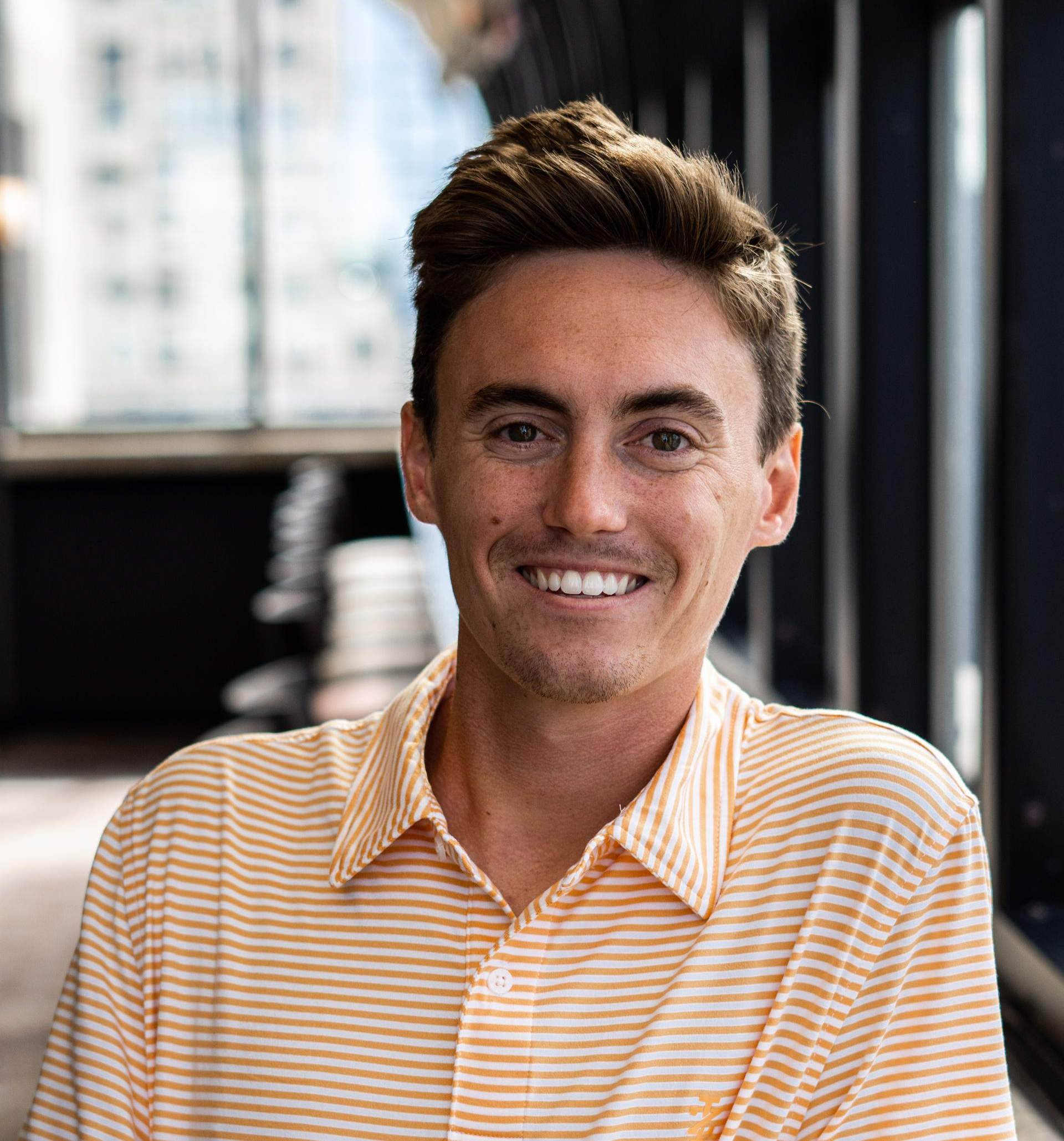 Man smiling, wearing a striped orange and white collared shirt, indoors near a window.