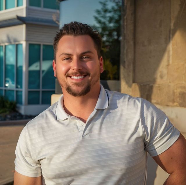 Man smiling in a light gray polo shirt, leaning against a stone structure, outdoor setting.