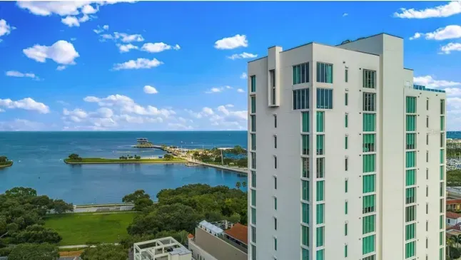 Modern white high-rise building with turquoise accents overlooks a bay and park under a blue sky.