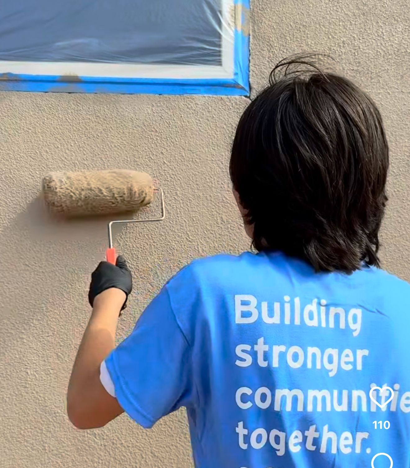 Person painting a wall with a roller. They are wearing a blue shirt that says 