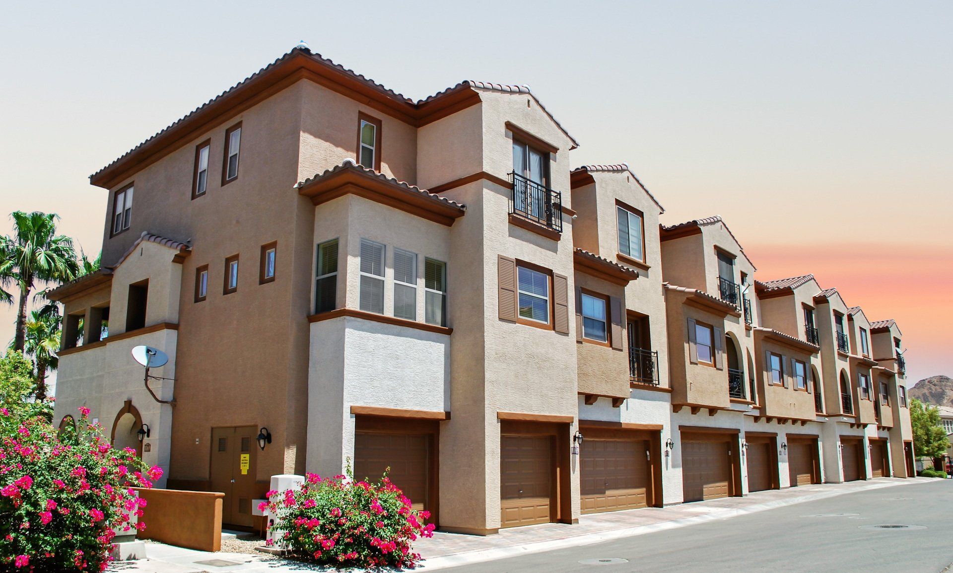 Row of townhouses, stucco exteriors, tan and white, with attached garages, in a sunny setting.
