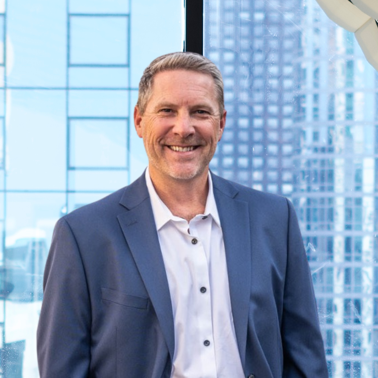 Man in a blue blazer smiles in front of a window, buildings in the background.