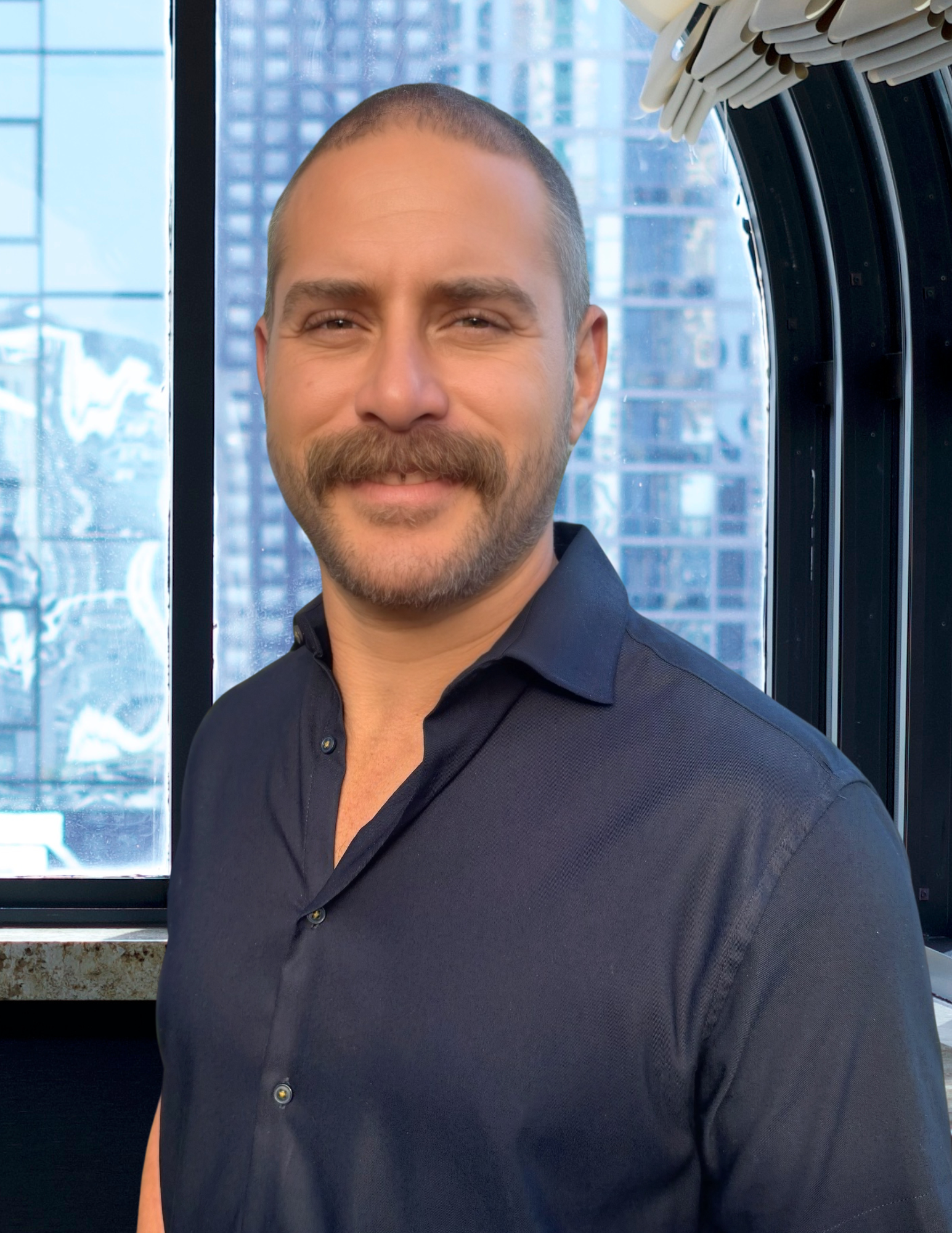 Man with dark hair and mustache wearing a dark blue shirt, smiling in front of a window.