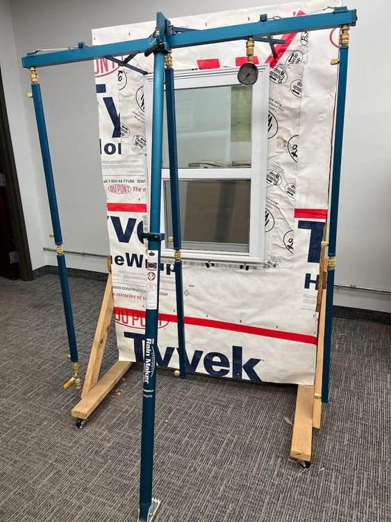 Blue scaffolding supporting a window frame covered in Tyvek wrap, set on a wooden base.