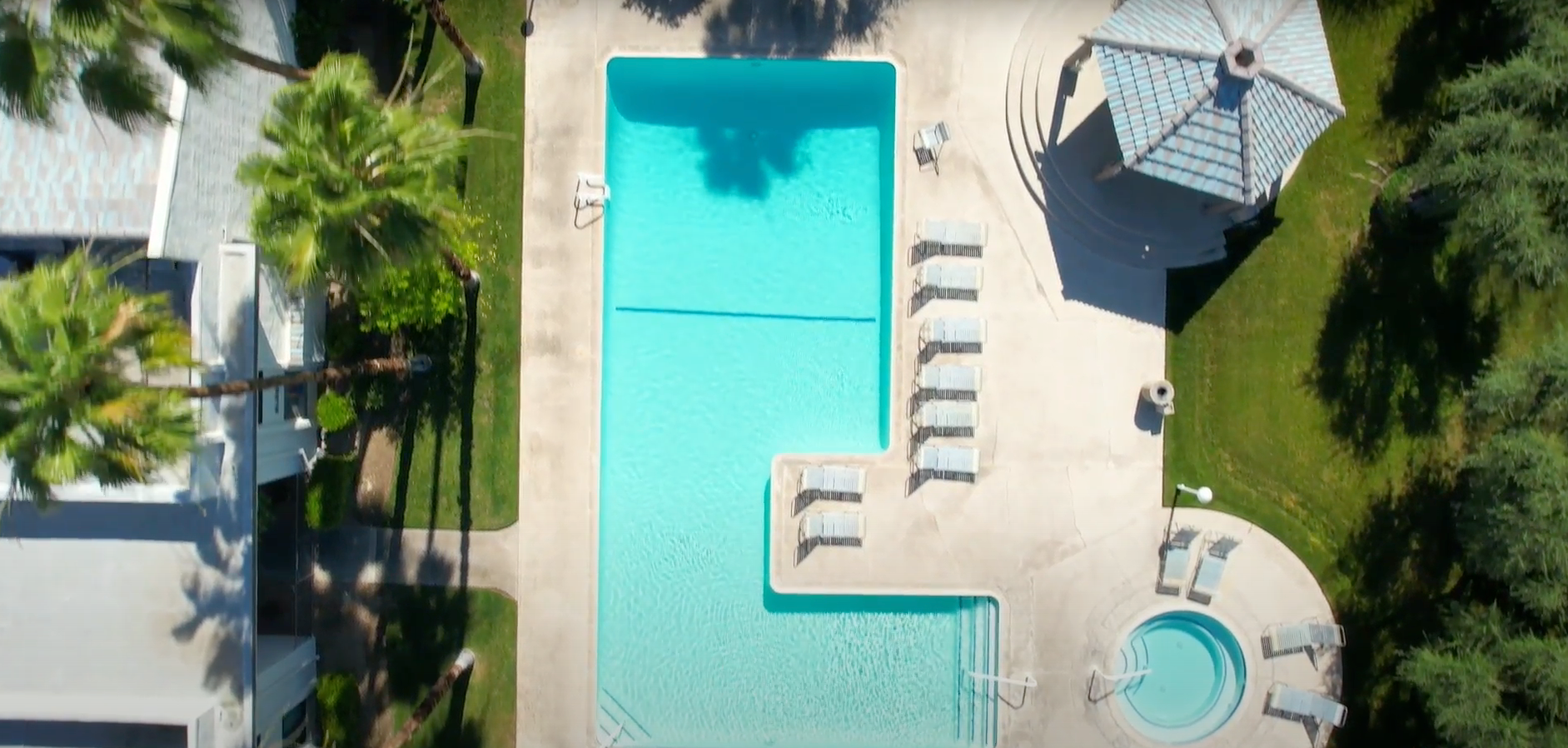 Aerial view of a rectangular swimming pool with a connecting, smaller pool. A gazebo and lounge chairs are nearby.