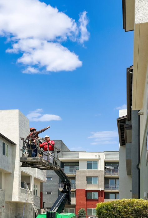 Workers on a crane lift near apartment buildings under a bright blue sky