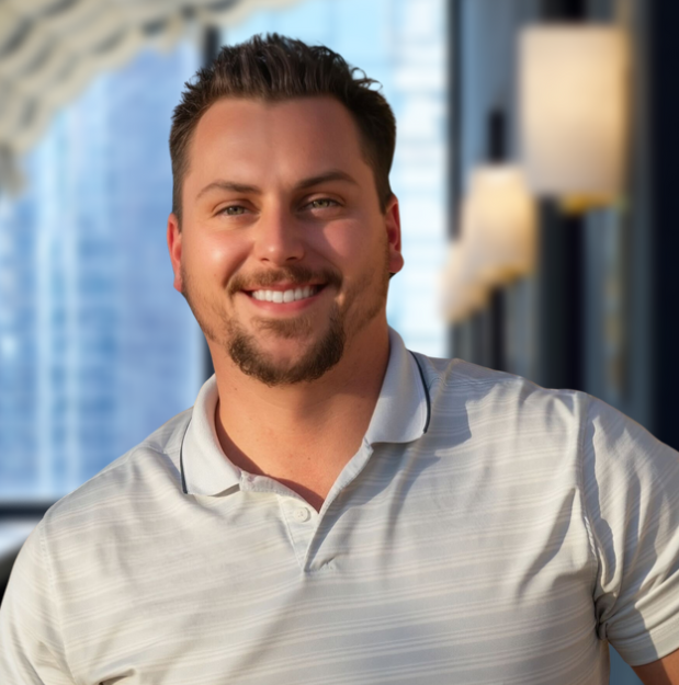 Man smiling, wearing a light gray polo shirt, in a modern interior with city view.