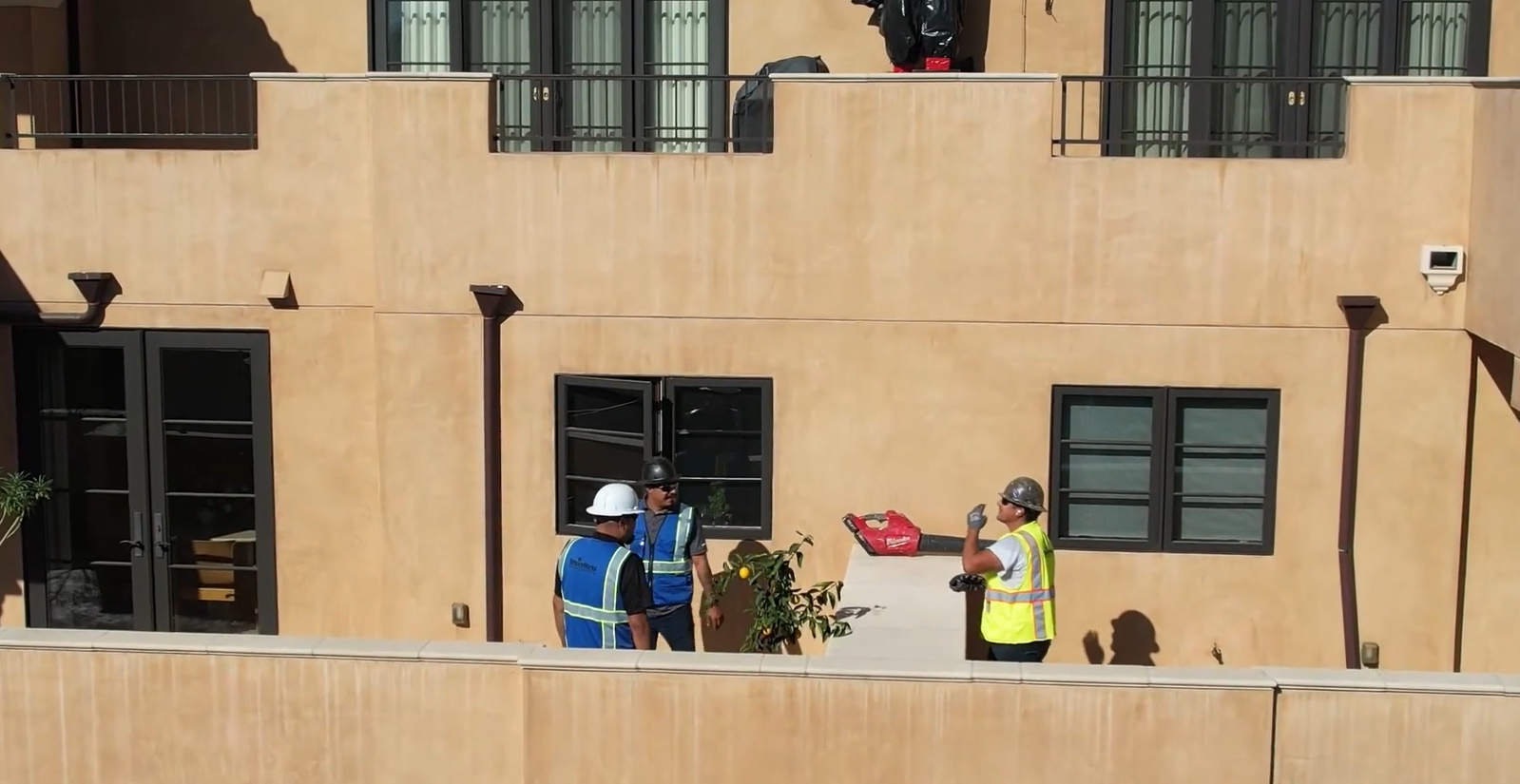 Three people on a balcony, one in a yellow safety vest holding flowers, against a tan apartment building.
