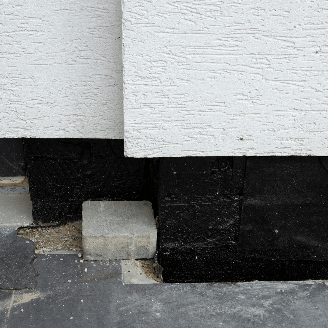 Close-up of a building's base: white textured wall, black waterproof coating, concrete block, and dark pavement.