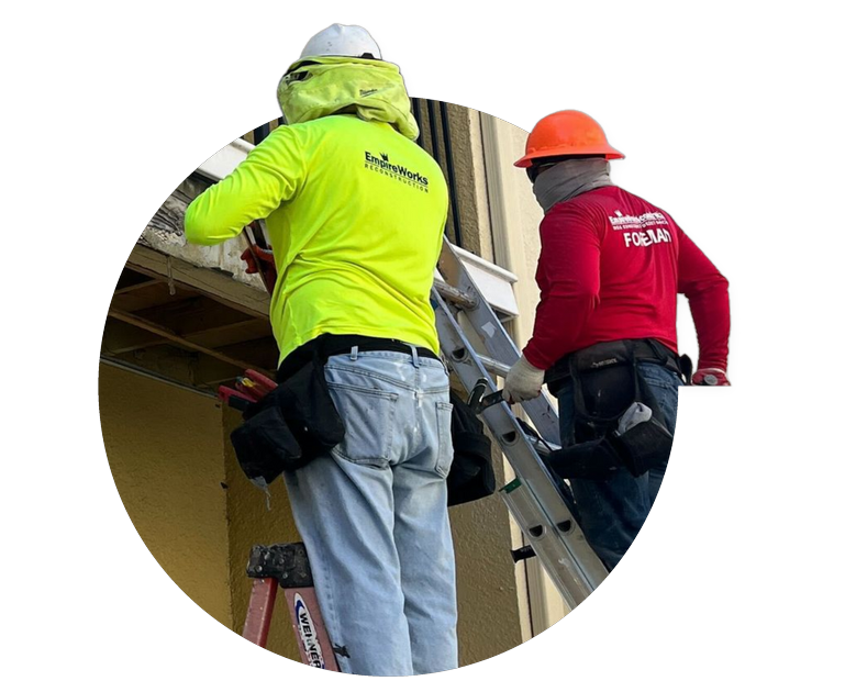 Two construction workers on a ladder inspecting a roof, wearing neon yellow and red jackets and hard hats