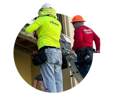 Two construction workers on a ladder inspecting a roof, wearing neon yellow and red jackets and hard hats