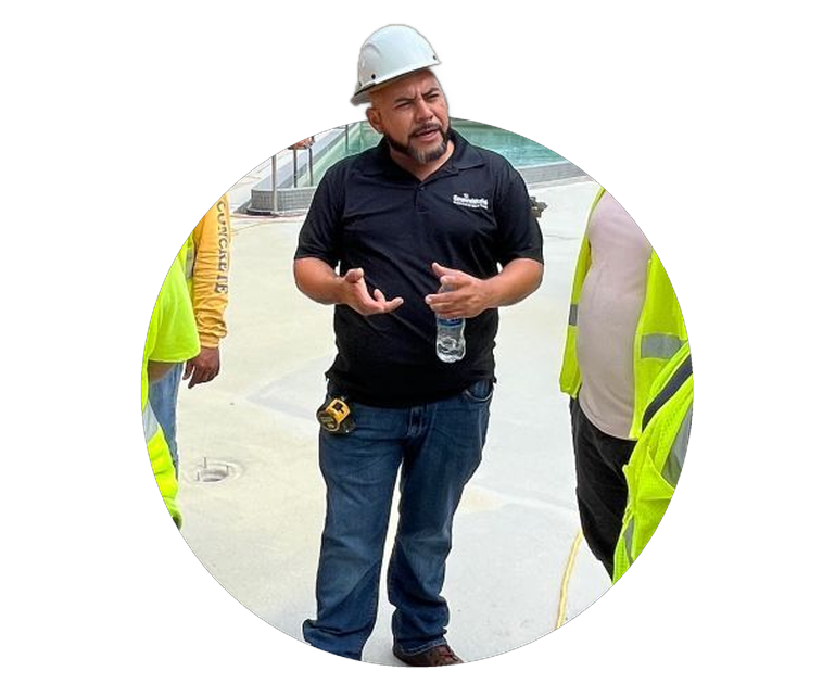 Man in hard hat and black polo speaking at a construction site, flanked by safety-vested workers.