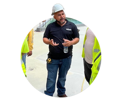 Man in hard hat and black polo speaking at a construction site, flanked by safety-vested workers.