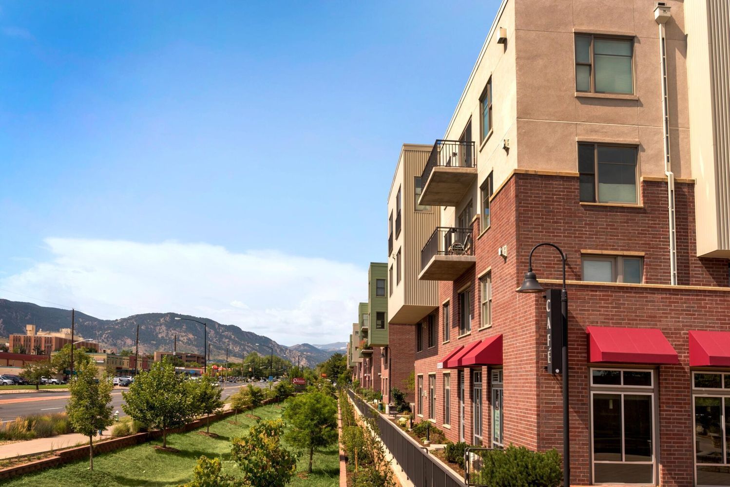 Apartment building with brick facade and red awnings, next to a green space with mountains in the background under a blue sky.