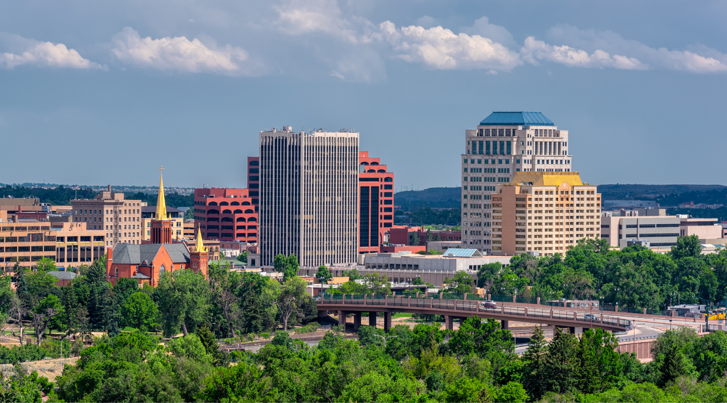 City skyline with buildings of varying heights and colors, under a blue sky with clouds.