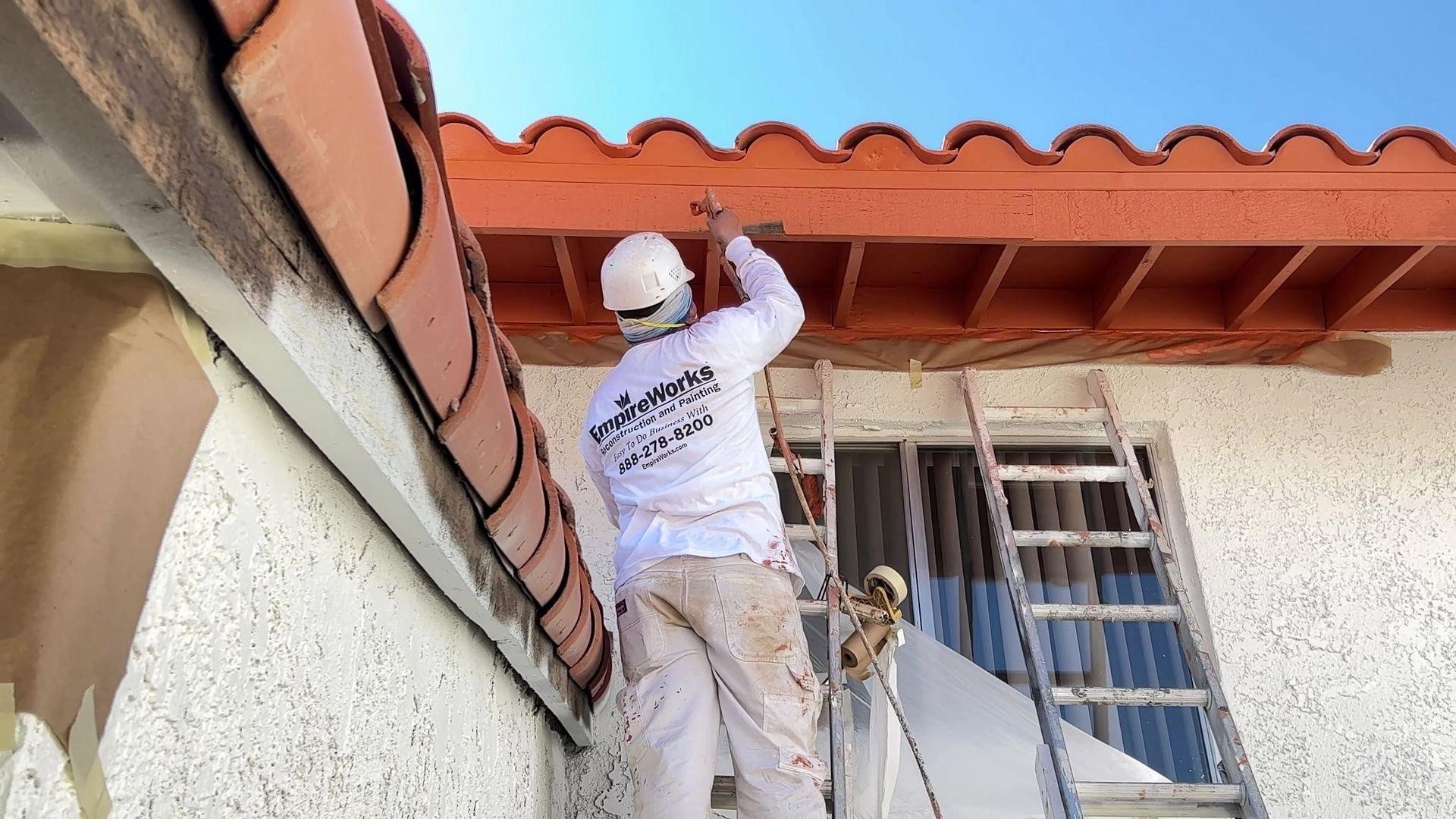Painter on a ladder, painting a roof's trim. Brown roof tiles and orange trim. Sunlit exterior.