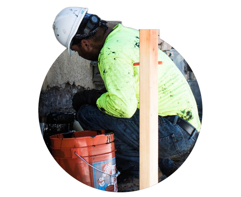 Worker in white hard hat and neon shirt crouches beside an orange bucket at a construction site.