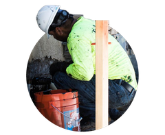 Worker in white hard hat and neon shirt crouches beside an orange bucket at a construction site.