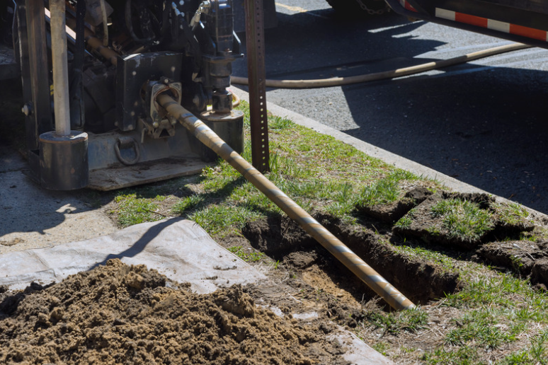 Drilling equipment boring into the ground next to a street. Dirt and turf are displaced.