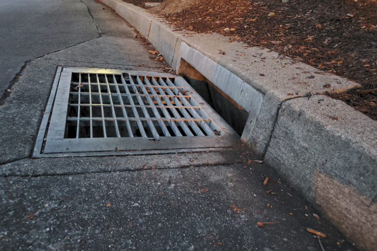 Metal drain in a concrete curb. Asphalt and dirt are visible.