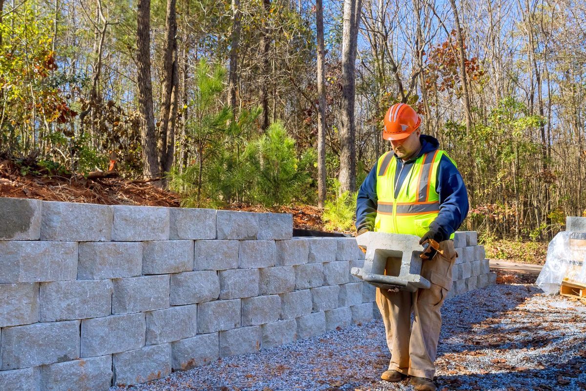 Construction worker near a stone retaining wall, holding a tool. He wears a reflective vest and hard hat, outdoors.