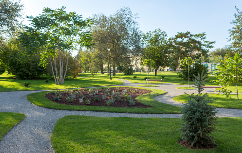 Park with curving gravel paths, green grass, trees, and a flower bed on a sunny day.