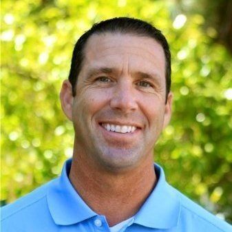 Man in blue shirt smiles, outside with blurred green background.
