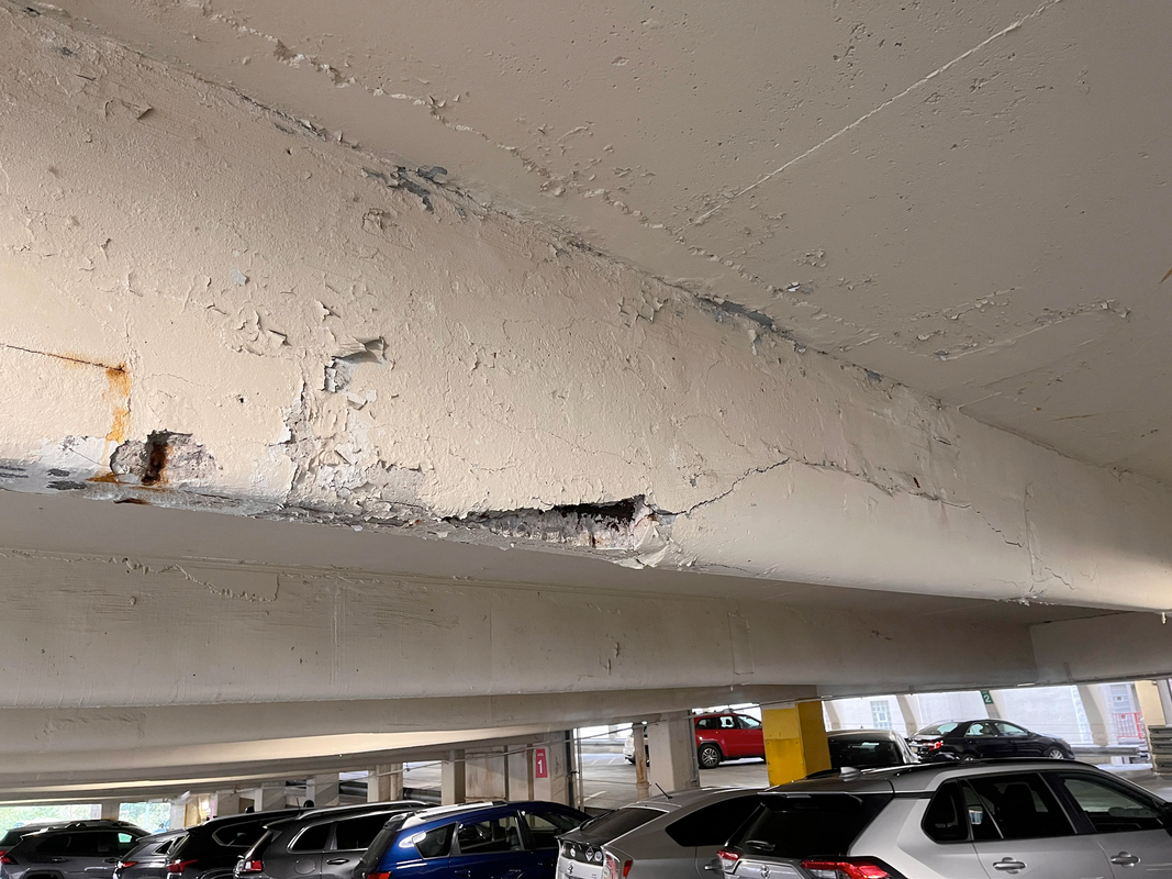 Overhead view of a concrete parking garage ceiling, showing cracks and exposed rebar, with parked cars below.