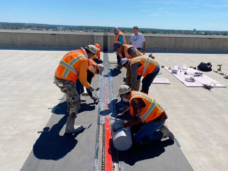 Construction workers repairing a roof expansion joint; they are wearing orange vests and hard hats.