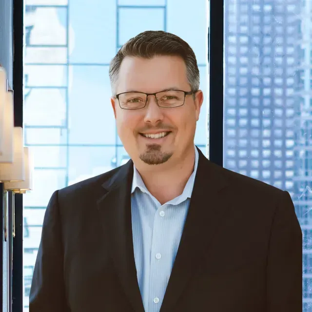 Man in a suit smiles, wearing glasses, in front of a window with a city skyline view.