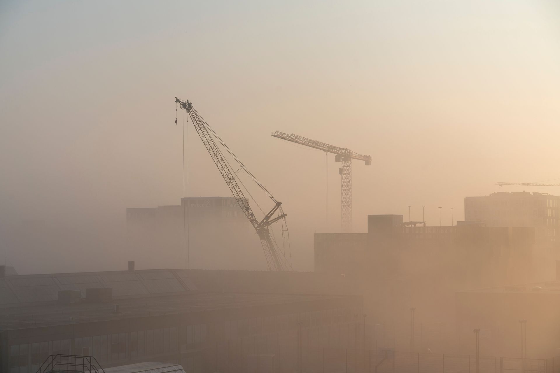 Two construction cranes silhouetted in a dusty, hazy city skyline at sunset